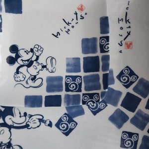 May include: Two white ceramic plates with blue and white Mickey Mouse designs. The larger plate features Mickey Mouse holding a paintbrush and the smaller plate features Mickey Mouse running. Both plates have the text "Mickey Mouse" in cursive.