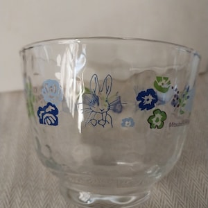 Puede incluir: Un cuenco de vidrio transparente con un diseño floral azul y verde con un conejo. El cuenco tiene una superficie texturizada y el texto "Mitsui Seito" es visible en la parte inferior.