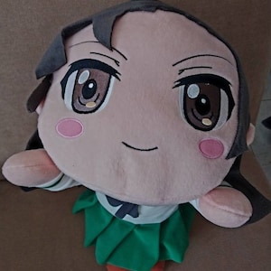 May include: A plush toy of a cartoon girl with brown hair, brown eyes, and a pink and green outfit. The toy is smiling and has a round face.