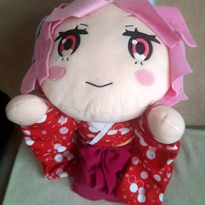 May include: A pink and red plush toy wearing a red and white patterned kimono. The toy has large eyes with red pupils and a small, smiling mouth.