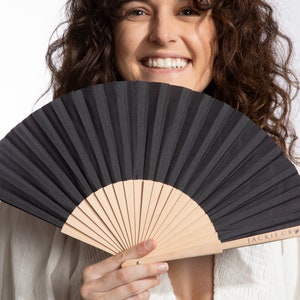 May include: A black hand fan with a light wood handle. The fan is open, displaying its pleated design. The handle has the text "JACKIE CRO" printed on it. The fan is held in front of a person.
