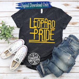 May include: A dark gray t-shirt with the words "LEOPARD PRIDE" in yellow outlined letters. The shirt is on a wooden surface with a pair of white sneakers and blue jeans.
