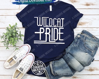 Pode incluir: Uma camiseta azul marinho com o texto "Wildcat Pride" em letras brancas. A camiseta está colocada sobre uma superfície de madeira com um par de tênis brancos e uma calça jeans azul.