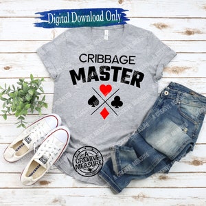 May include: A gray t-shirt with the text "Cribbage Master" in black. The shirt also features a graphic of four playing card suits: a heart, a spade, a diamond, and a club.