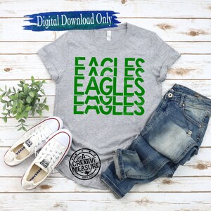 May include: A gray t-shirt with the word "EAGLES" repeated four times in green letters. The shirt is laid out on a wooden surface with a pair of white sneakers and blue jeans.