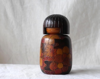 May include: A wooden Kokeshi doll featuring a dark brown, rounded head with a smooth, straight-cut bob hairstyle. The cylindrical body is decorated with intricate, hand-painted floral patterns in shades of deep red, orange, and yellow.