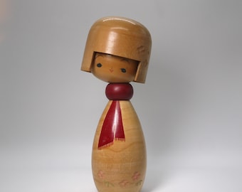 May include: A wooden Kokeshi doll with a light brown head and body. The doll has a red scarf and a small, smiling face. The body is decorated with pink floral designs.