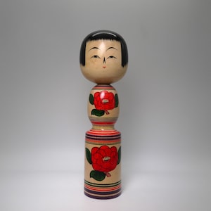 May include: A wooden Kokeshi doll with a smooth, rounded head featuring painted black hair and a serene facial expression. The cylindrical body is decorated with red camellia flowers, green leaves, and horizontal stripes in red and purple.