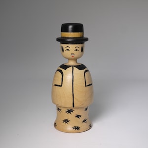 Könnte beinhalten: Eine Holzfigur einer Person mit schwarzem Zylinderhut mit gelbem Band, einer schwarz umrandeten Jacke und einem Rock, der mit schwarzen sternartigen Formen verziert ist. Das Gesicht hat bemalte Merkmale, darunter einen roten Mund.