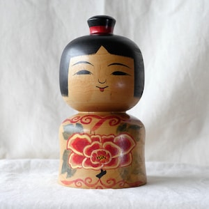 May include: A wooden Kokeshi doll featuring a rounded head with black painted hair and a red top knot. The body is cylindrical with a natural wood finish, decorated with a large red and yellow floral design and green leaves at the base.