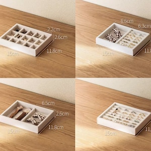 May include: Four white jewelry organizer trays with various internal compartment configurations. Each tray measures 15 cm by 11.8 cm by 2.6 cm. The trays feature dividers for rings, watches, and small accessories, with some sections showing specific dimensions like 2.2 cm, 6.6 cm, 6.3 cm, and 6.5 cm.