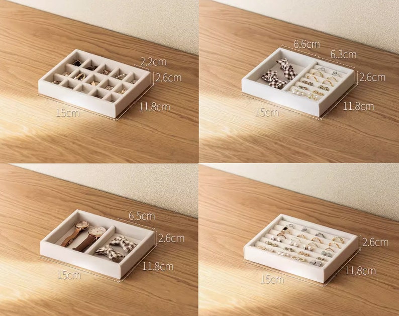 May include: Four white jewelry organizer trays with various internal compartment configurations. Each tray measures 15 cm by 11.8 cm by 2.6 cm. The trays feature dividers for rings, watches, and small accessories, with some sections showing specific dimensions like 2.2 cm, 6.6 cm, 6.3 cm, and 6.5 cm.
