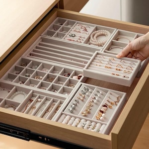 May include: A light beige velvet jewelry organizer tray set with multiple compartments, ring slots, and earring holders, arranged inside a wooden drawer. The modular trays contain various rings, necklaces, bracelets, and earrings.
