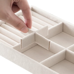 May include: A rectangular beige jewelry organizer tray with a soft velvet-like finish. The tray features multiple compartments, including a section with padded ring rolls and adjustable dividers that can be repositioned to customize the storage space.