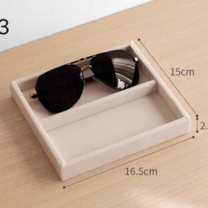 May include: A beige velvet-lined square storage tray with a divider, holding black sunglasses. The tray is labeled A3 and measures 16.5 cm in length, 15 cm in width, and 2.5 cm in height. The interior is divided into two rectangular sections.