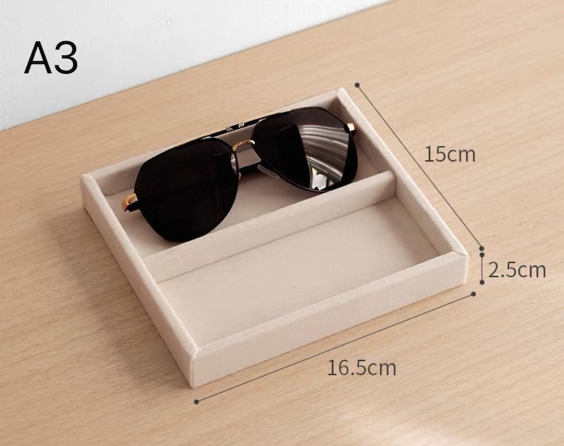 May include: A beige velvet-lined square storage tray with a divider, holding black sunglasses. The tray is labeled A3 and measures 16.5 cm in length, 15 cm in width, and 2.5 cm in height. The interior is divided into two rectangular sections.