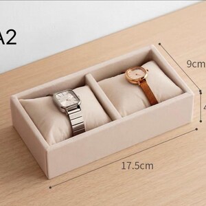 May include: A beige velvet watch display tray labeled A2 with two compartments, each containing a soft cushion. One cushion holds a silver-tone watch with a metal link band, and the other holds a gold-tone watch. Dimensions are 17.5cm by 9cm by 4.5cm.