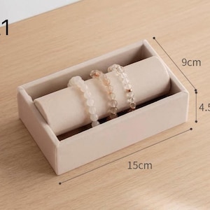 May include: A beige velvet jewelry display tray labeled A1, featuring a cylindrical holder with three beaded bracelets. The rectangular tray measures 15 cm in length, 9 cm in width, and 4.5 cm in height, providing a structured base for accessories.