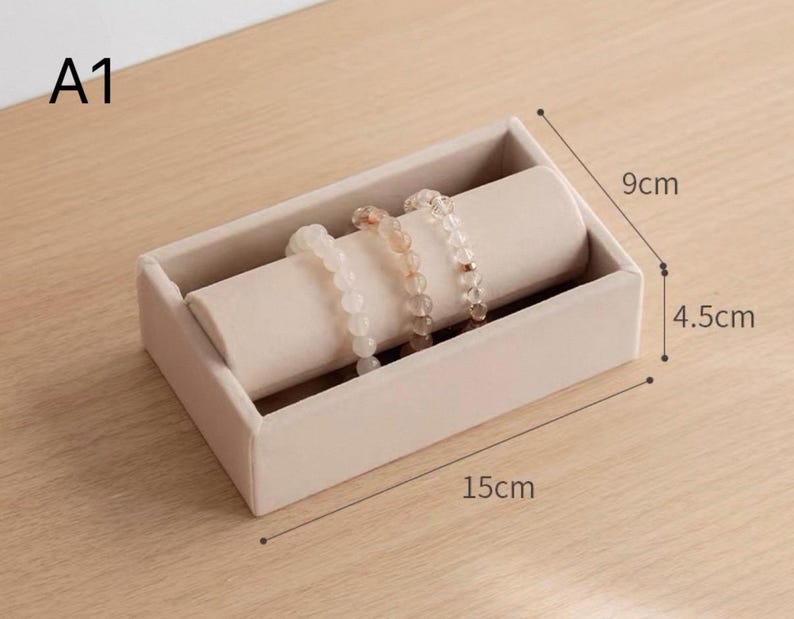 May include: A beige velvet jewelry display tray labeled A1, featuring a cylindrical holder with three beaded bracelets. The rectangular tray measures 15 cm in length, 9 cm in width, and 4.5 cm in height, providing a structured base for accessories.