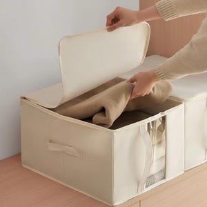 Puede incluir: Una caja de almacenamiento rectangular de tela beige con tapa abatible y una ventana de plástico transparente en el panel frontal. La caja cuenta con asas laterales y contiene un artículo textil color canela doblado en su interior.