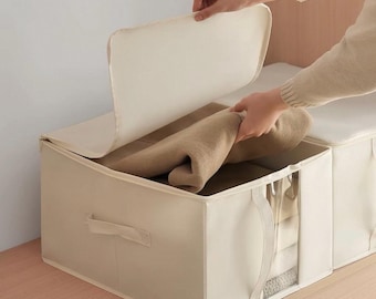 May include: A rectangular beige fabric storage bin with a hinged lid and a clear plastic window on the front panel. The bin features side handles and contains a folded tan textile item, providing a structured solution for organizing household items.