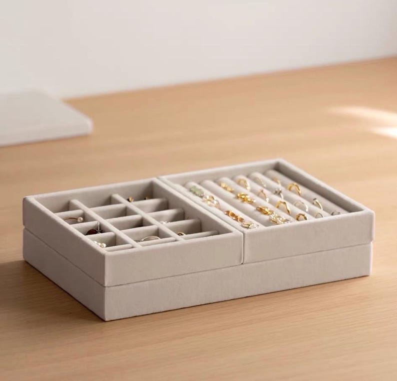 May include: A light gray velvet jewelry organizer tray featuring two distinct sections. The left side contains a grid of square compartments, while the right side features multiple padded slots designed to hold rings and small earrings securely.