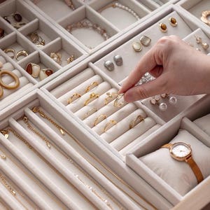 May include: A beige velvet jewelry organizer tray featuring multiple compartments filled with gold and silver rings, earrings, necklaces, and a wristwatch on a small cushion. The tray is organized with rows for rings and chains.