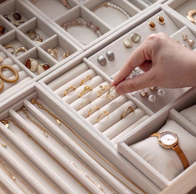 May include: A beige velvet jewelry organizer tray featuring multiple compartments filled with gold and silver rings, earrings, necklaces, and a wristwatch on a small cushion. The tray is organized with rows for rings and chains.
