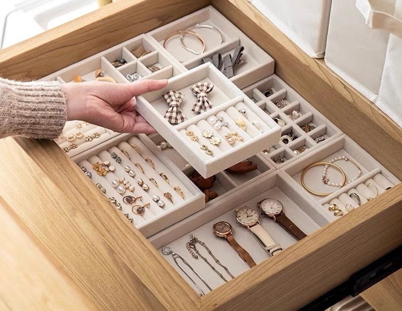 May include: A set of modular beige velvet jewelry organizer trays arranged inside a wooden drawer. The trays feature various compartments, including ring rolls and open sections, holding earrings, necklaces, bracelets, watches, and hair bows.