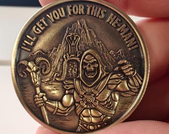 May include: Bronze-colored coin featuring a detailed relief of a skeletal figure with a hood and armor, holding a staff. The coin's edge has the inscription "I'LL GET YOU FOR THIS HE-MAN!" with a mountain and cobra design in the background.