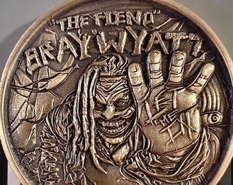May include: A bronze-colored coin featuring a detailed relief of a character with the text "THE FIEND" and "BRAY WYATT". The character has a sinister grin and outstretched hand. The coin has a textured, aged appearance.