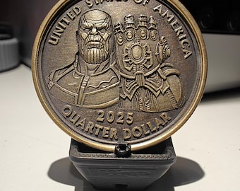 May include: A bronze-colored quarter dollar coin featuring a detailed relief of a character with a gauntlet. The coin is inscribed with "UNITED STATES OF AMERICA" and "QUARTER DOLLAR 2025". The coin is displayed on a black stand.