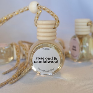May include: A clear glass bottle with a wooden cap and a white label that reads "Perisa rose oud & sandalwood car diffuser | 8ml". The bottle is hanging from a gold and white rope.