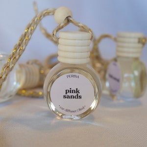 May include: A clear glass car diffuser with a white label that says "Perisa pink sands car diffuser | 8ml". The diffuser is hanging from a gold and white string.