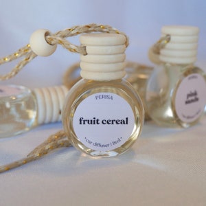 May include: A clear glass car diffuser bottle with a white wooden cap and a gold string. The bottle has a white label with the text "Perisa fruit cereal . car diffuser | 8ml."