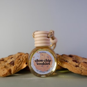 May include: A small glass bottle with a wooden cap and a rope hanger. The bottle is filled with a light yellow liquid and has a label that says "choc chip cookies" and "car diffuser 8ml". The bottle is surrounded by two chocolate chip cookies.