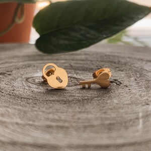 May include: Gold-coloured stud earrings with a lock and key design. The lock earring has a keyhole, and the key earring has a heart-shaped head. The earrings are displayed on a textured, grey surface.