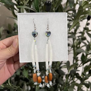 May include: A pair of dangle earrings with heart-shaped iridescent abalone shells. The earrings feature white, elongated beads, clear crystal accents, and brown wooden beads. The earrings are displayed on a white background.