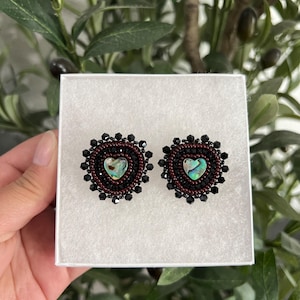 May include: A pair of heart-shaped earrings. Each earring features a heart-shaped abalone shell center, surrounded by a dark red beaded border and black beads. The earrings are presented in a white box.