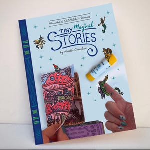 May include: A light blue book titled "Tiny Magical Stories" by Arielle Crenshaw. The cover features illustrations of a gnome, a mermaid, and a dragon. A glue stick and a miniature paper house are also visible.