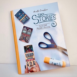 May include: A book titled "Tiny Stories: Cut & Fold Matchbox Dioramas" by Arielle Crenshaw. The book cover features illustrations of miniature houses and a pair of scissors.