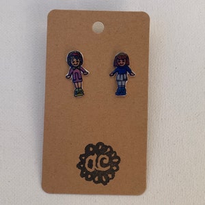 May include: A pair of stud earrings featuring two cartoon girls with brown hair. One girl is wearing a pink shirt and the other is wearing a blue shirt. The earrings are on a brown card with a black circle logo that says "ac".