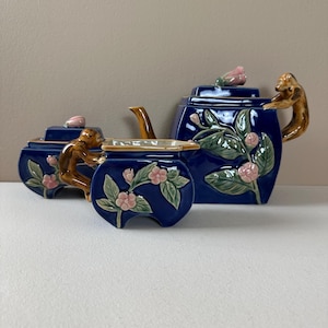 May include: A dark blue ceramic tea set consisting of a teapot, a lidded sugar bowl, and a creamer. Each piece features sculpted brown monkeys, raised green leaves, and light pink flower buds. The teapot lid is topped with a pink flower bud.