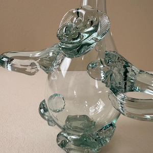 May include: A clear glass decanter featuring a sculpted monkey figure integrated into the body. The monkey has raised arms forming handles on each side, with a detailed face, textured ears, and a rounded torso. The glass is transparent and smooth.