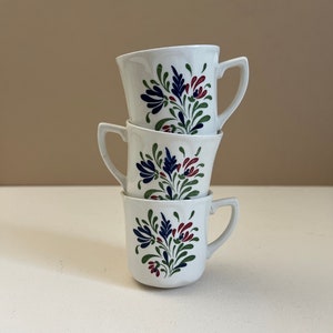 Puede incluir: Tres tazas de café de cerámica blanca con un diseño floral en tonos azules, verdes y rojos. Las tazas están apiladas una encima de la otra.