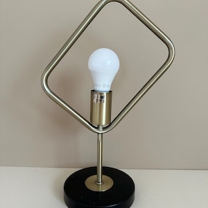Peut inclure: Une lampe de table moderne au design géométrique. La lampe comprend une ampoule blanche dans une douille couleur laiton, soutenue par une fine tige métallique et une base circulaire noire. Un cadre carré entoure l'ampoule. La lampe a un cordon d'alimentation marron.