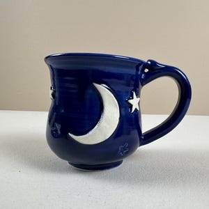 May include: A deep blue ceramic mug featuring a glossy finish with a prominent white crescent moon and several small white star accents embossed on the exterior surface. The mug has a curved handle and a rounded base, showcasing a celestial design.