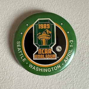 Puede incluir: Un botón verde y negro con un borde dorado. El botón presenta un gráfico del Space Needle en Seattle, Washington. El texto "1989 NCAA FINAL FOUR" está impreso en el botón. El texto "SEATTLE, WASHINGTON, APRIL 1-3" está impreso alrededor del borde.