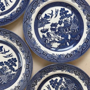 May include: A collection of round ceramic plates featuring a classic blue and white willow pattern. Each plate displays a detailed transfer print of a traditional landscape with pagodas, willow trees, a bridge, and birds in flight against a cream background.
