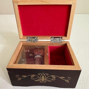 May include: Open wooden music box with a dark brown exterior and a light brown frame. The interior is lined with red velvet. The box features a glass-covered compartment with visible musical components and a decorative gold floral design.